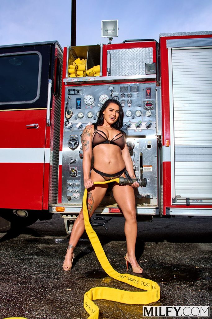 Busty female firefighter posing in sheer lingerie and holding a big hose by a fire truck