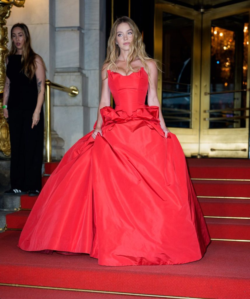 Sydney Sweeney heading down the steps of a hotel staircase and wearing a big red dress