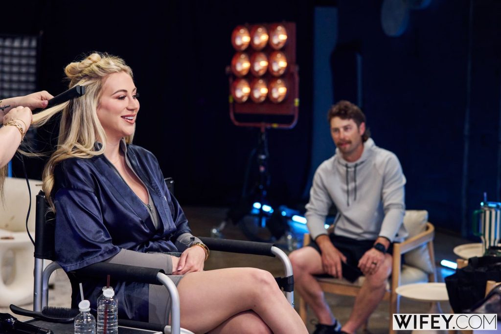 Blonde wife wearing a robe while sitting in the makeup chair and her husband is looking on