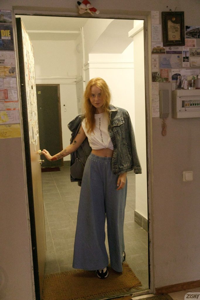 Redhead posing in the hallways of an apartment building wearing loose pants and a jacket