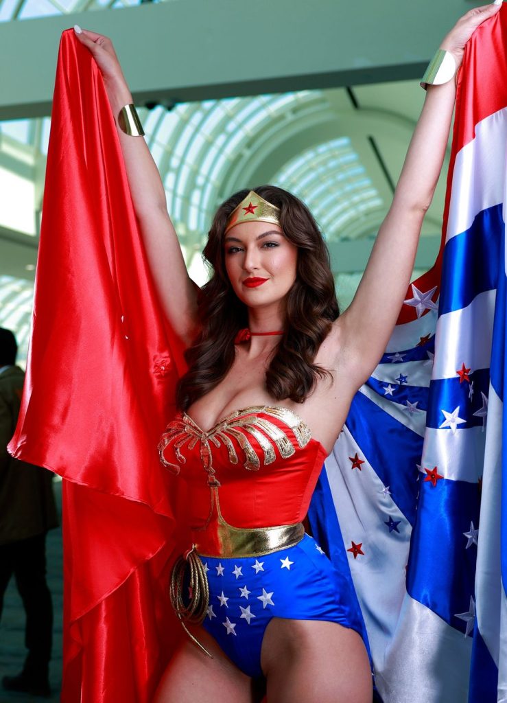 Hot brunette dressed as Wonder Woman lift her arms and cape in the air at Comic Con