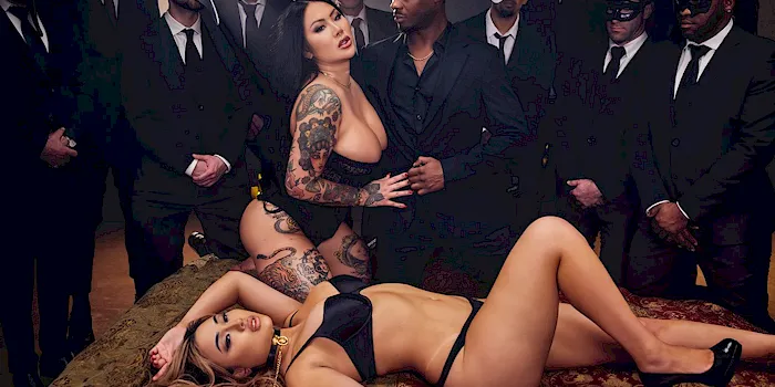 Curvy and busty Asian porn stars in lingerie in the middle of a circle of men watching