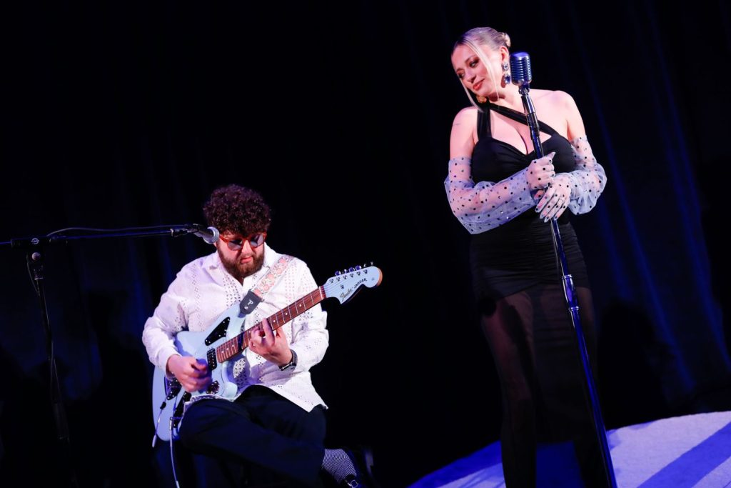 Beautiful blonde singer looking at the guitar player while they perform on stage with moody blue lighting