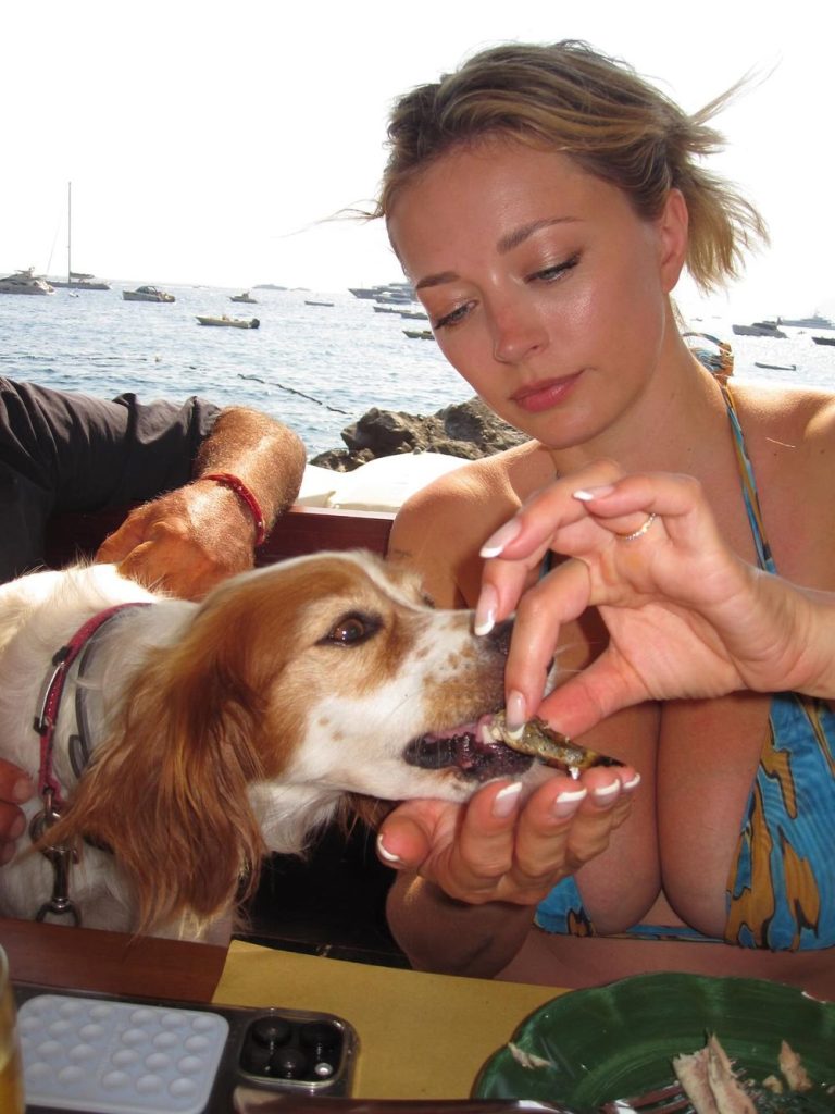 Blonde babe in a bikini top giving a dog some food while sitting at a restaurant