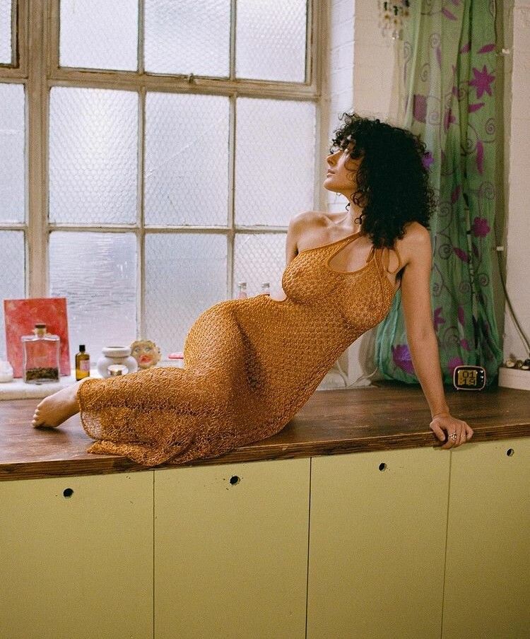 Sexy brunette nude under a sheer orange dress while she sits on her side on top of the counter in a retro kitchen
