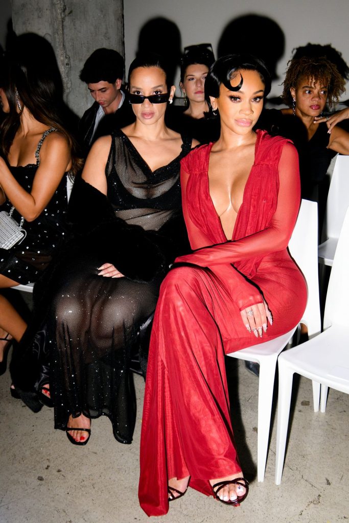 Far away view of Saweetie next to a woman in black sitting down on white chairs at a show