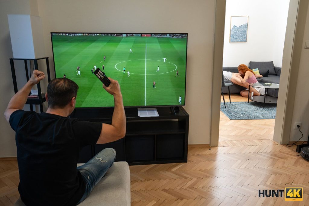 Man cheering on a Soccer match on TV while a girl is performing oral sex on a man in the other room