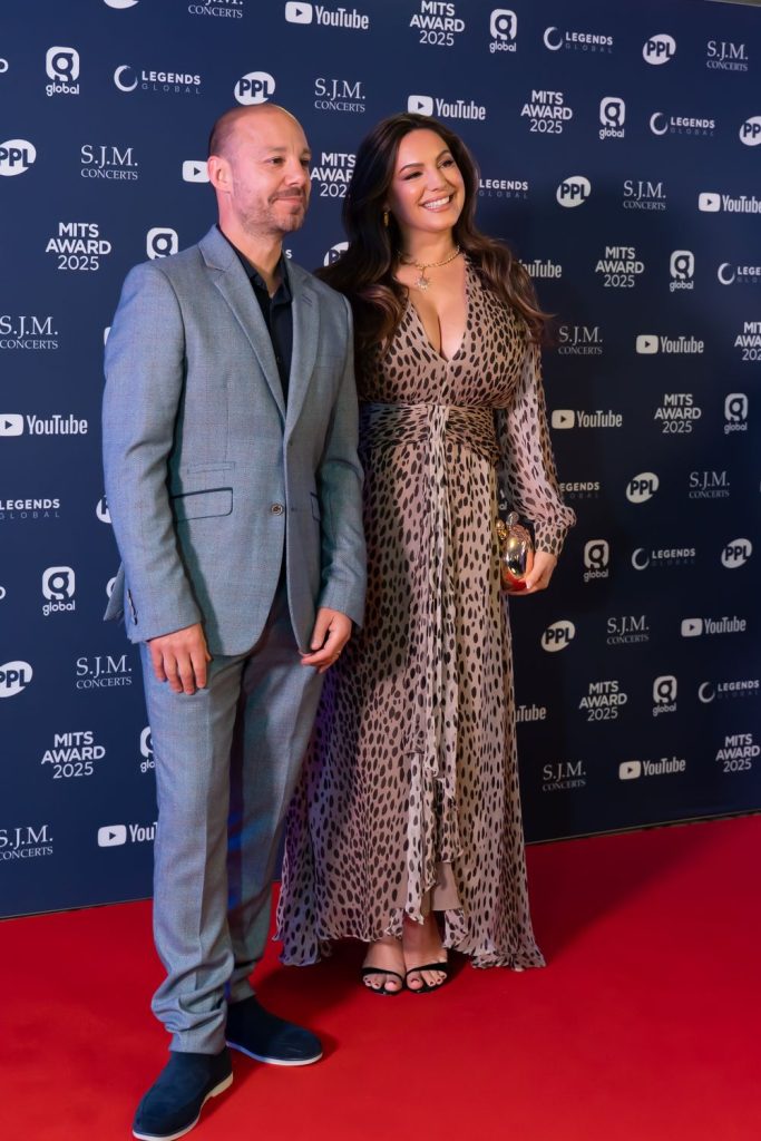 Full body view of Kelly Brook posing next to a man on the red carpet at a slight angle