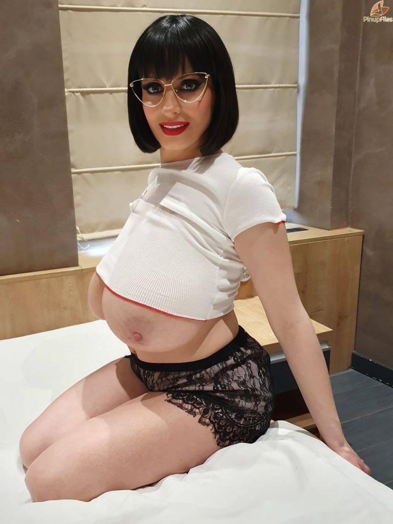 Full body side view of woman sitting on her knees in bed with top pulled up and the bottom half of her large breasts are visible