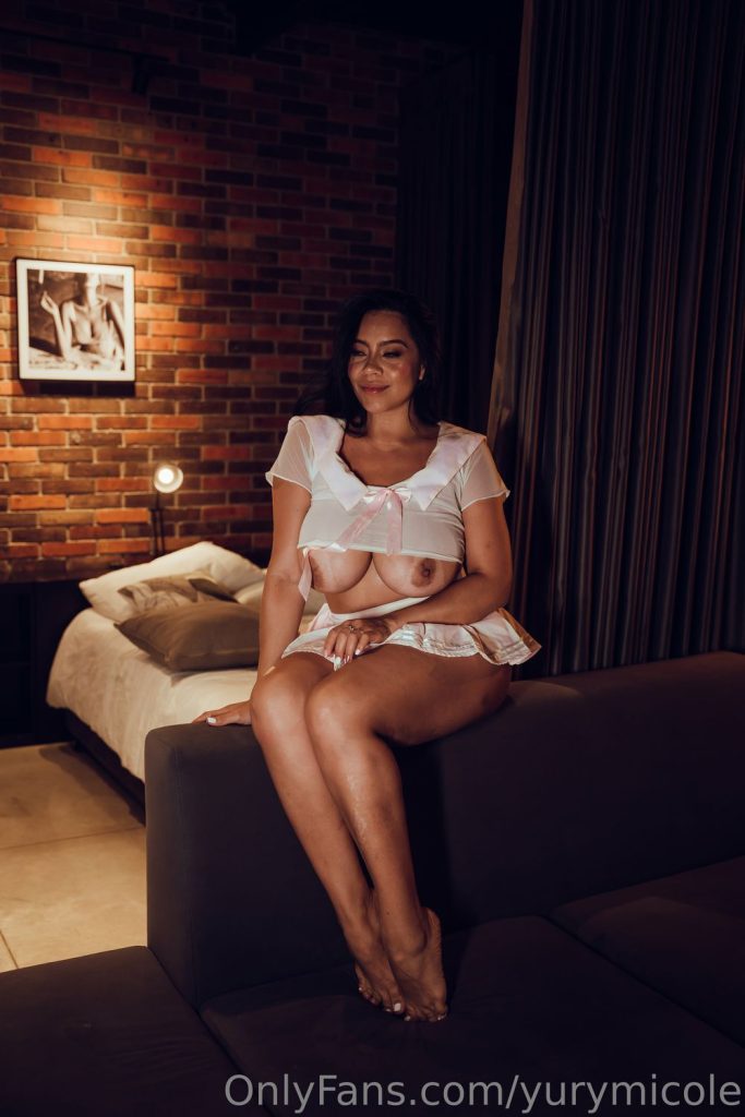 Full body front view of Latina girl sitting on top of a sofa with breasts hanging out of her top