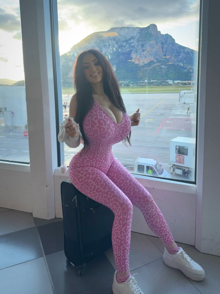 Full body frontal view of woman with large breasts sitting on top of a suitcase inside an airport