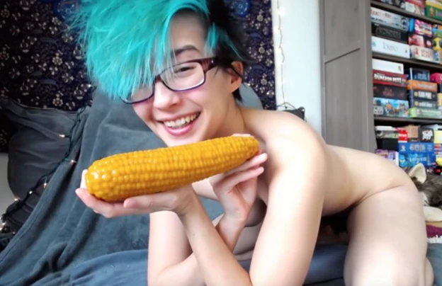 Nude girl on all four facing the camera and smiling while holding a dildo shaped like a corn on the cob