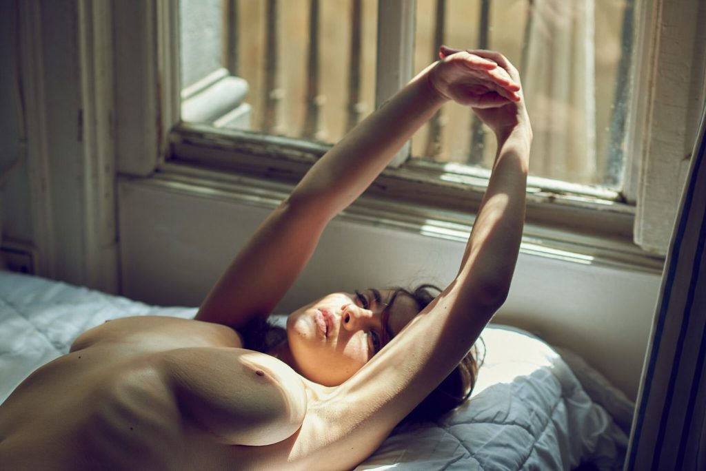 Close up of a topless woman stretching her arms above her head as she lays in bed and the sunlight hitting her face