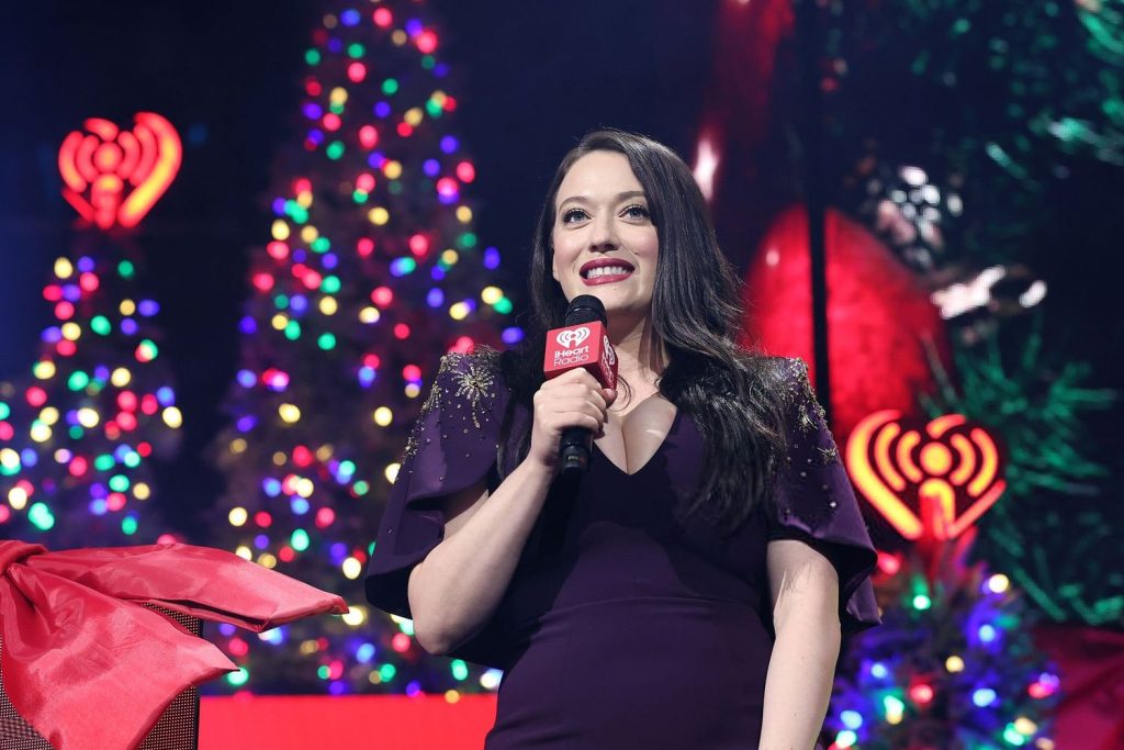 Close up of Kat Dennings smiling and holing a microphone in front of Christmas trees