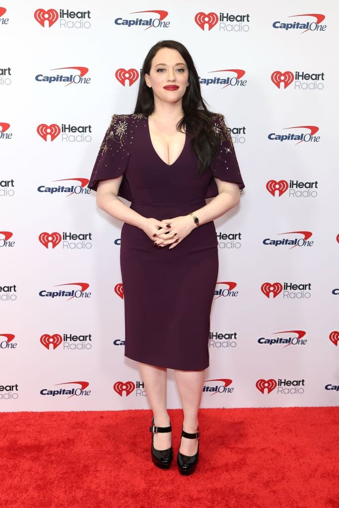 Frontal view of Kat Dennings posing on a red carpet with both hands clasped in front of her