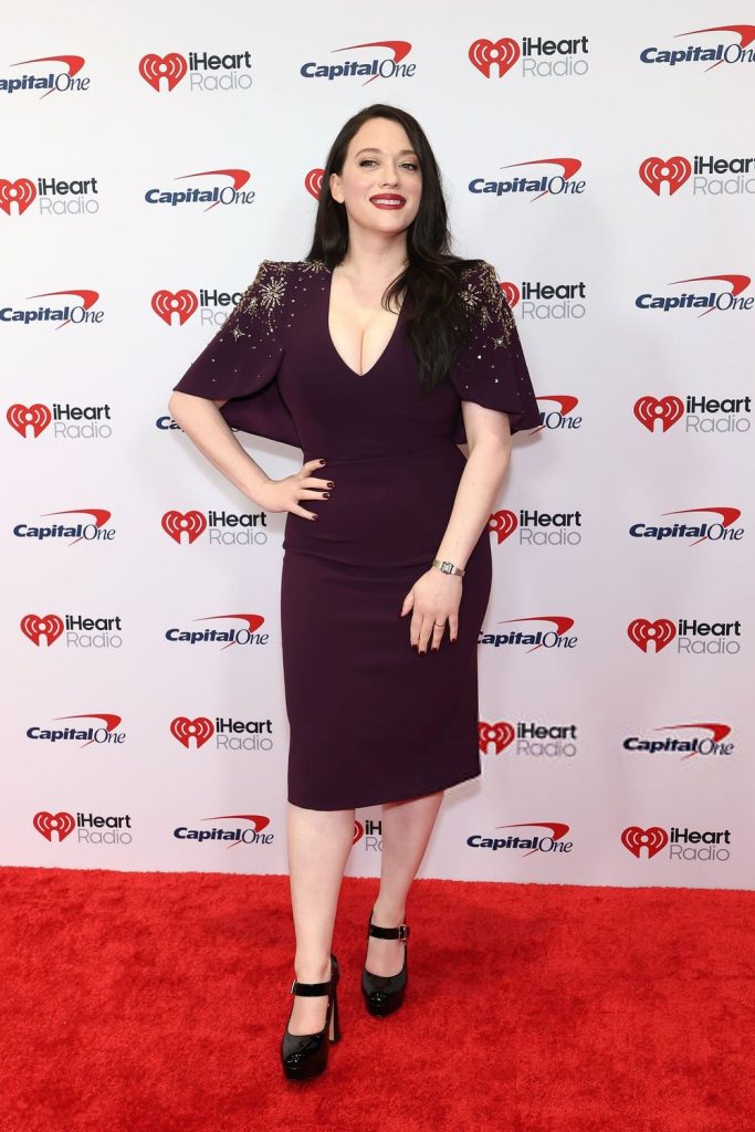 Frontal view of Kat Dennings posing on a red carpet with the other hand on her hip