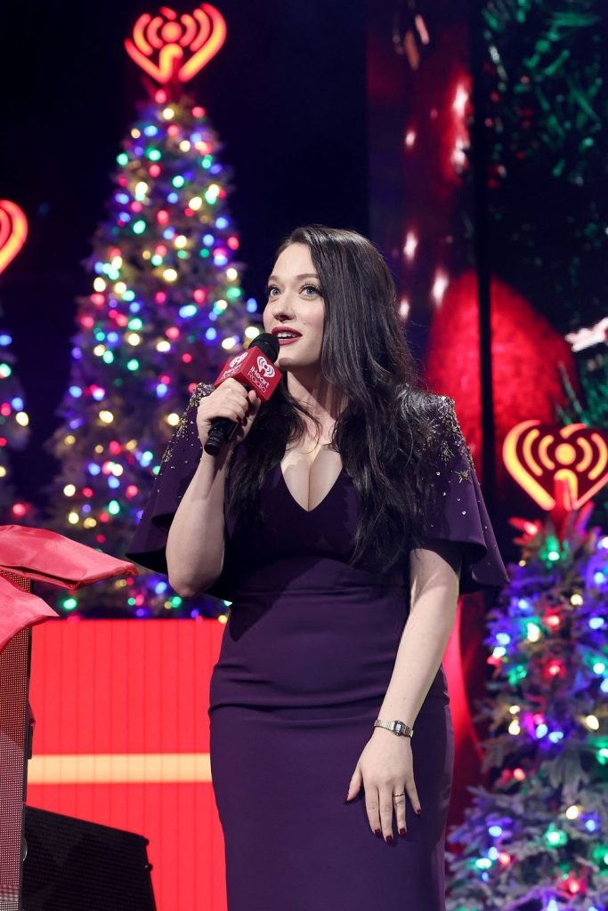 Frontal of Kat Dennings talking into a microphone in front of Christmas trees
