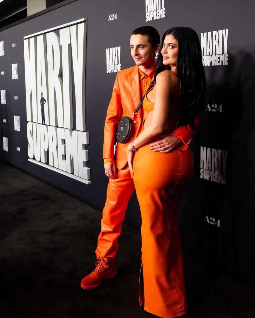Side and full body view of Kylie Jenner turning her back towards the camera while posing next to Timothée Chalamet on a red carpet next to each other in matching outfits