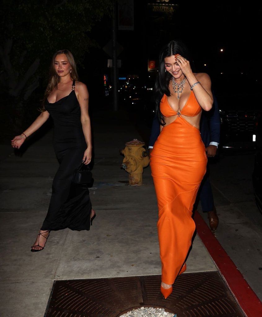Far away frontal view of Kylie Jenner looking down, touching her head while walking along a sidewalk and with a woman in the background