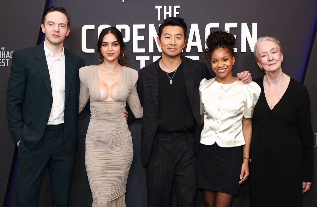 Frontal of two men and three women posing on a red carpet next to each other