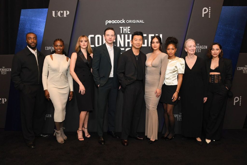 Far away frontal of two men and five women posing on a red carpet next to each other