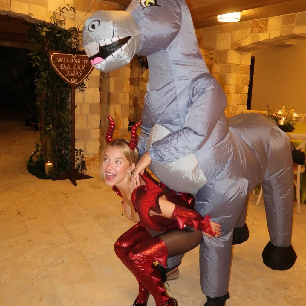 Sydney Sweeney laughing while wearing a dragon costume and carrying a giant donkey on her back