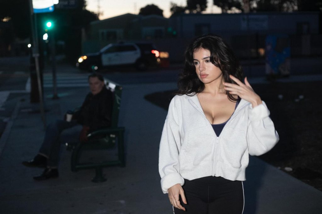 Outdoors at dusk with a woman walking towards the camera and a man watching her from a bench in the background