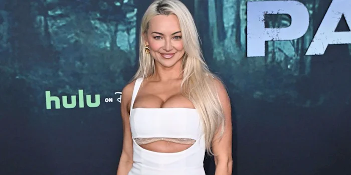 A woman with long blonde hair in a white dress poses and smiles on a red carpet, with a Hulu logo and a forest-themed background behind her.