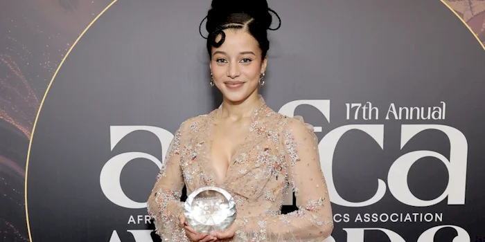 Chase Infinity in a gown and holds a glass award and smiles at the camera against a backdrop with large white letters.
