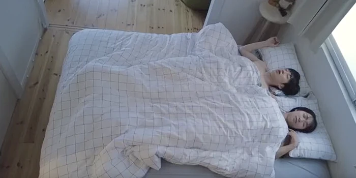 Asian couple are lying side by side in bed, covered with a white checkered blanket, with light coming through a nearby window. The bedroom has wooden floors and a minimalistic style.