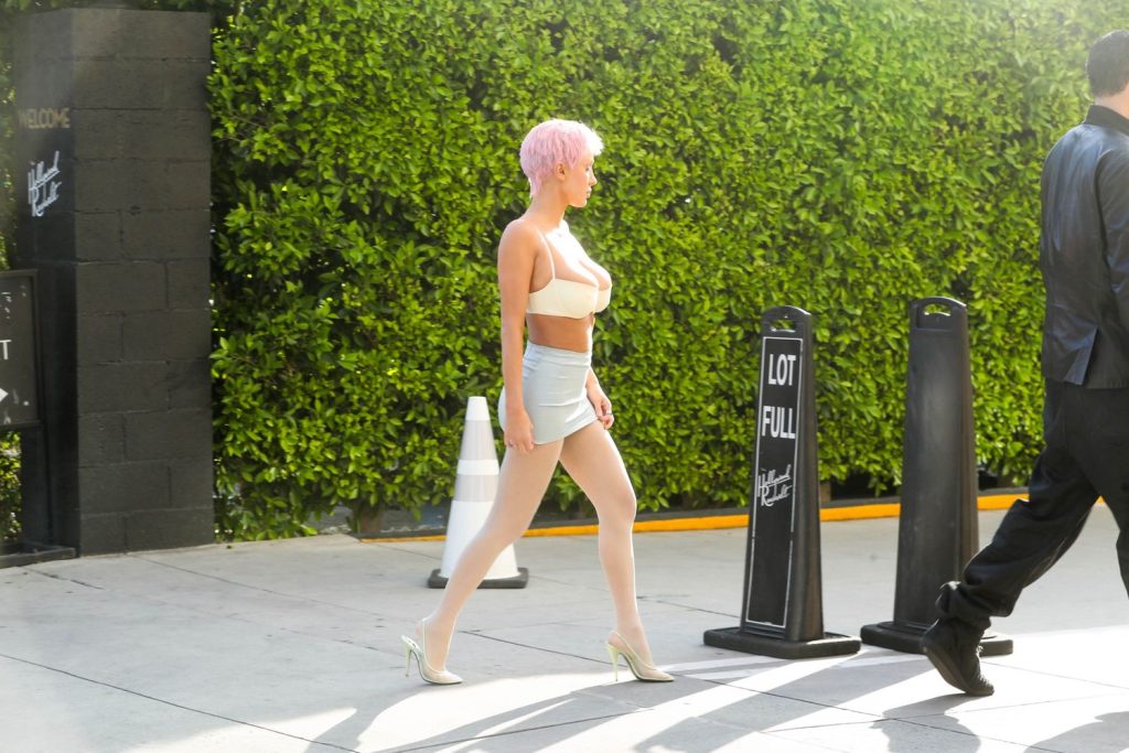 A woman with short pink hair, wearing a beige crop top, light blue mini skirt, white stockings, and heels walks on a sidewalk past Lot Full signs and a green hedge.
