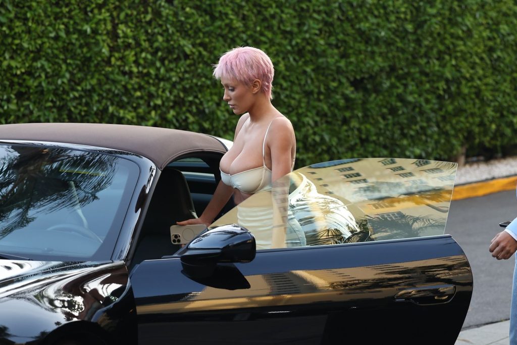 A woman with short pink hair wearing a white dress getting into a black car, holding a smartphone. Green hedges and a building are visible in the background.
