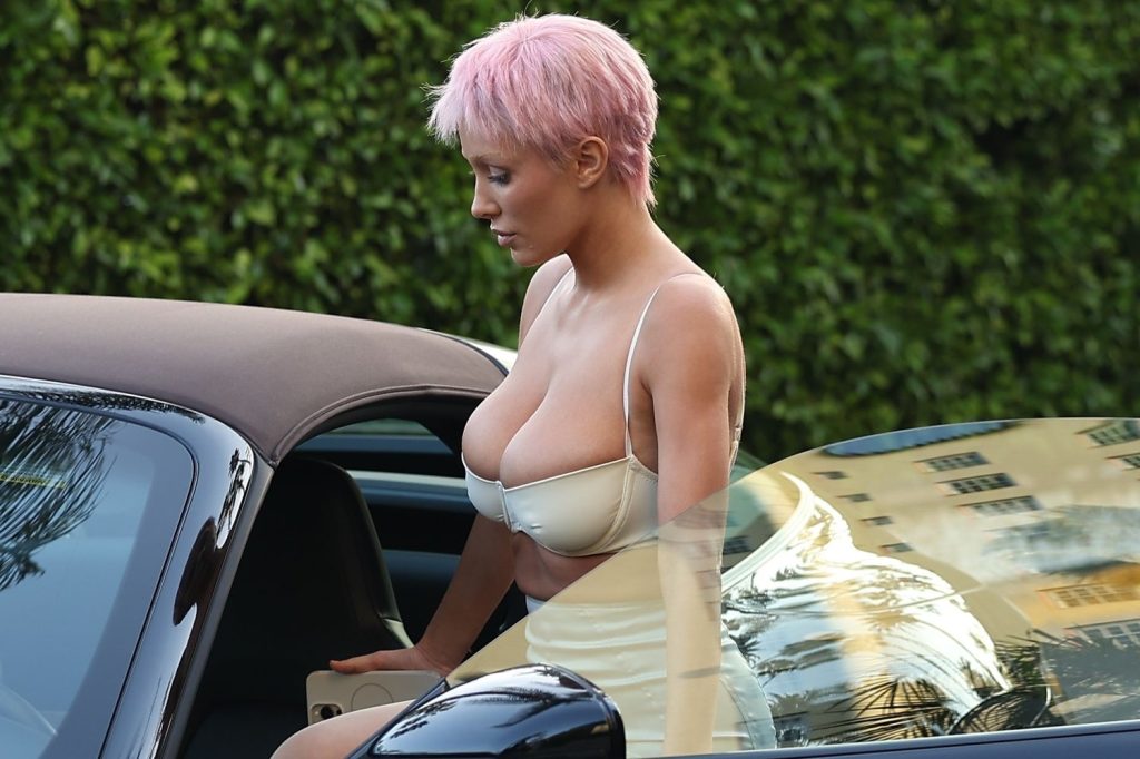 Close up of a woman with short pink hair wearing a cream-colored bra top steps out of a black convertible car. Green shrubbery is visible in the background.