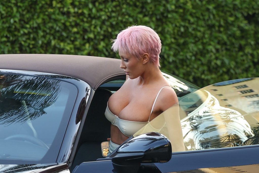 A woman with short pink hair and a cream-colored top leans down as she gets into a black car with a tan roof, with greenery in the background.