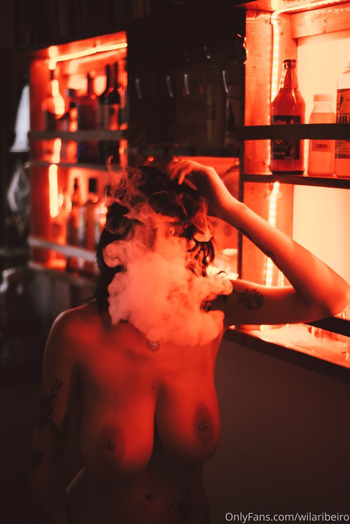 A topless woman with smoke coming out of her mouth lean against a bar