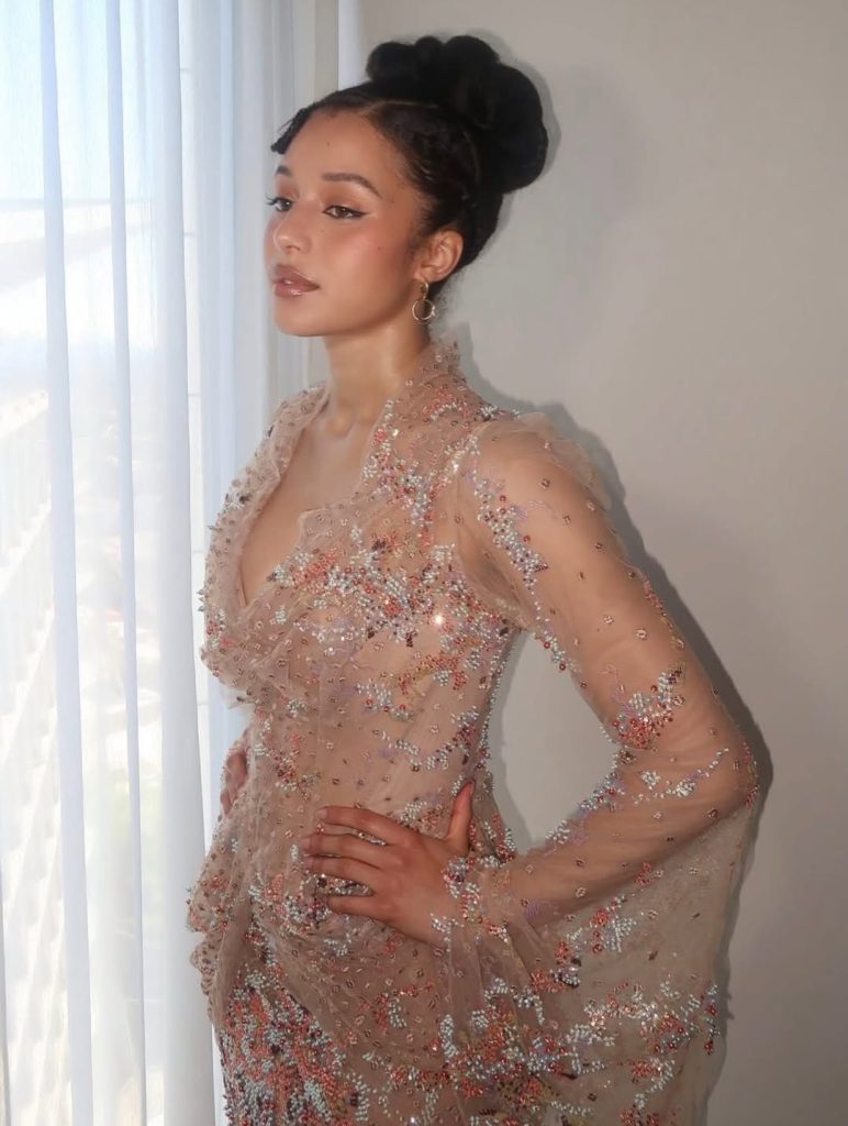 A woman stands by a window in natural light, wearing an elegant, sheer, beaded gown with floral details. Her dark hair is styled in an updo, and she gazes calmly to the side with one hand on her hip.