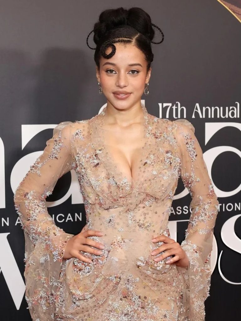A woman poses on the red carpet in a sheer, embellished gown with a deep neckline and flared sleeves. She has styled hair, subtle makeup, and stands in front of a backdrop with text from an awards event.