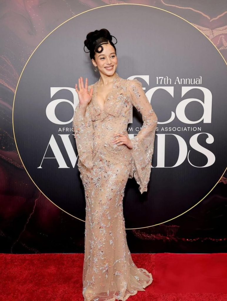 A woman in a sheer, embellished gown waves as she poses on a red carpet in front of a backdrop reading 17th Annual AFRICAN FILM CRITICS ASSOCIATION AWARDS.