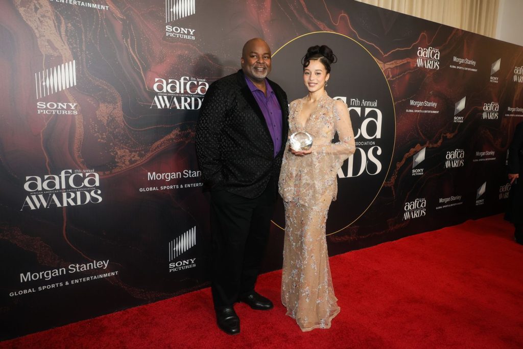 A man in a black suit and a woman in a sparkling beige gown holding a glass award pose together on the red carpet at the 7th Annual AAFCA Awards, with event logos in the background.