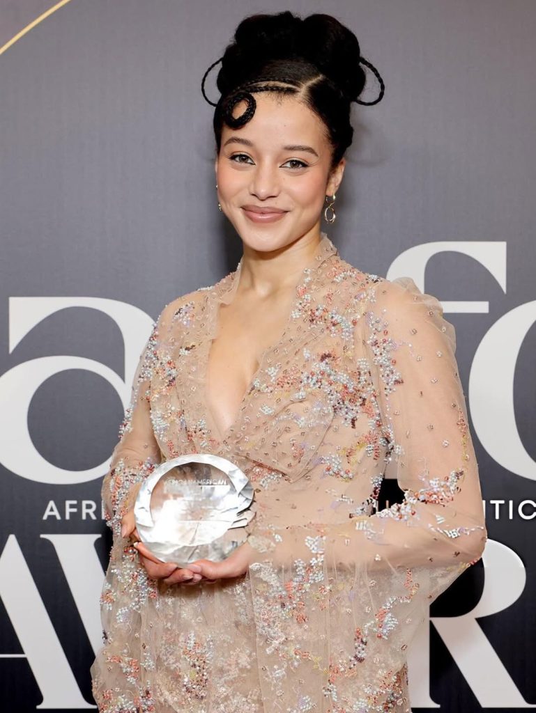 A woman in a sheer, embellished gown holds a glass award and smiles at the camera against a backdrop with large white letters.