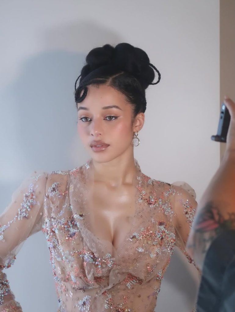 A woman with an elegant updo hairstyle poses against a plain background, wearing a sheer, beaded dress with floral embellishments. She gazes to the side while someone holds a camera in the foreground.