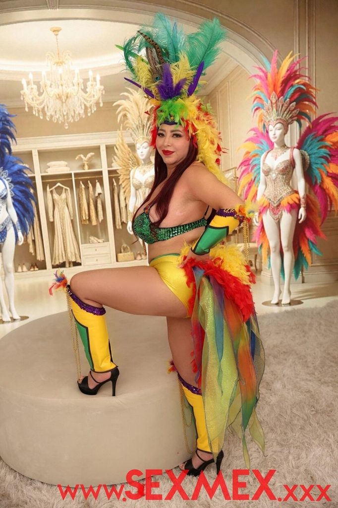 A woman in a colorful carnival costume with feathers poses indoors on a white circular platform. Behind her, mannequins in elaborate feathered outfits and a closet with dresses are visible.