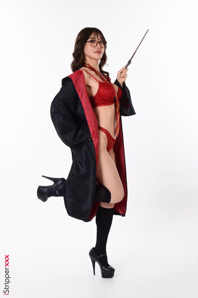 A woman poses in a red bra and underwear set, a black robe with red lining, a striped tie, black knee-high socks, and high heels. She holds a wand and stands on one leg against a plain white background.