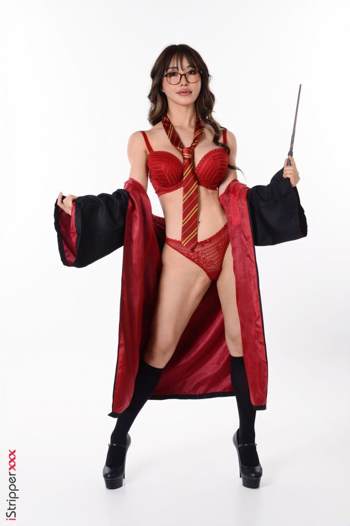 A woman dressed in red lingerie, a Gryffindor-style necktie, black robe, black knee-high socks, and heels holds a wand and stands against a white background.