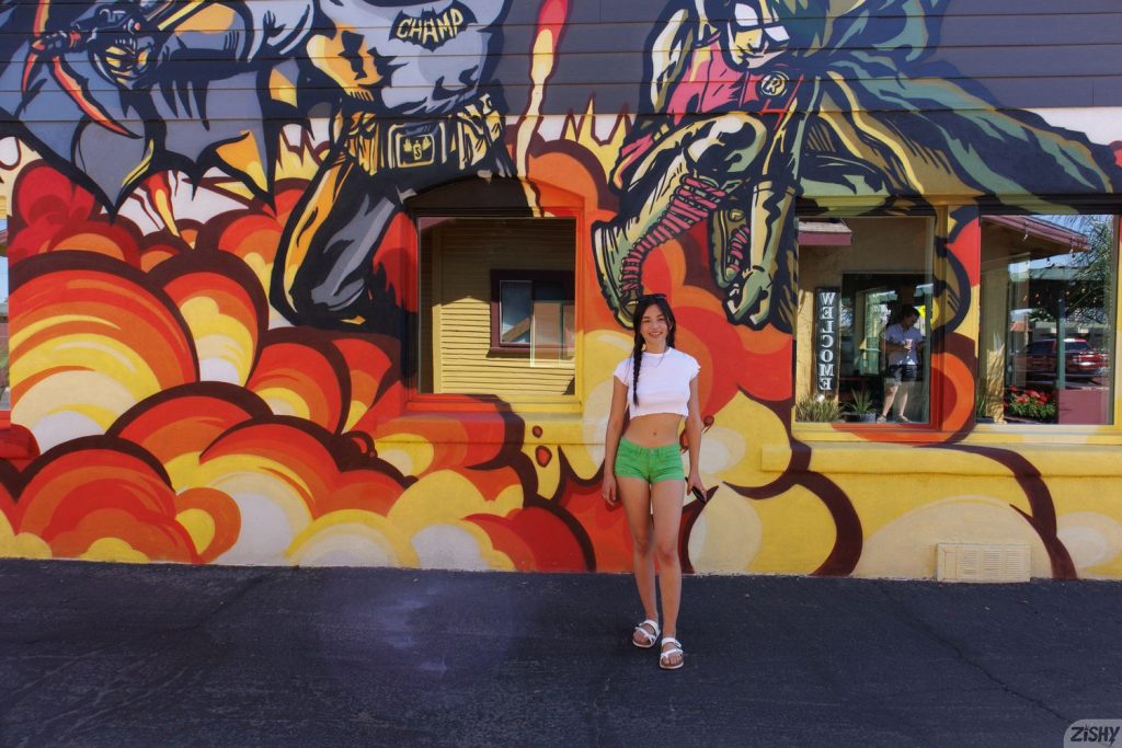 A smiling woman in a white crop top and green shorts stands in front of a colorful comic book-style mural featuring action scenes and bold graphics.