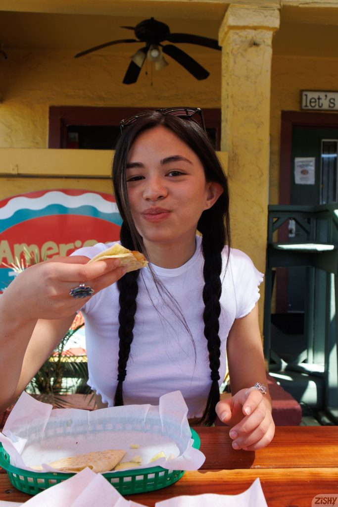 A young woman with long dark braids and a white t-shirt sits at an outdoor table, smiling while holding a tortilla chip. There is a green basket with food in front of her and a colorful wall in the background.