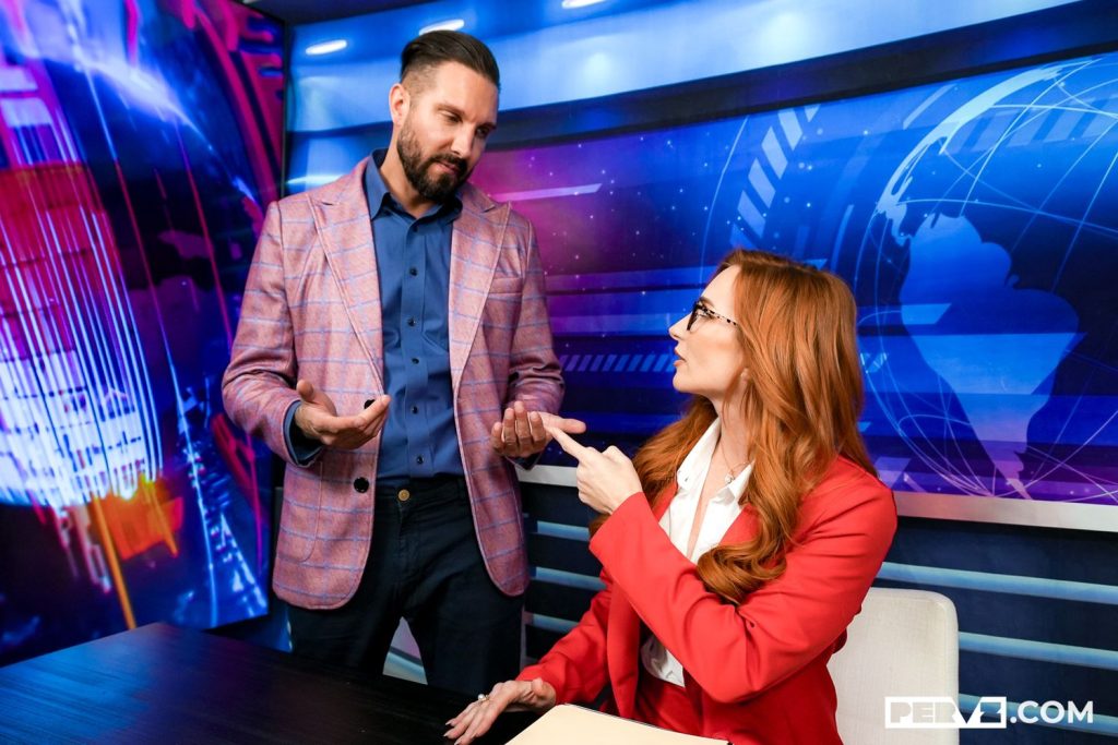 A man in a plaid blazer stands talking to a seated woman in a red blazer, both engaged in discussion in a modern newsroom set with digital graphics and a world map in the background.
