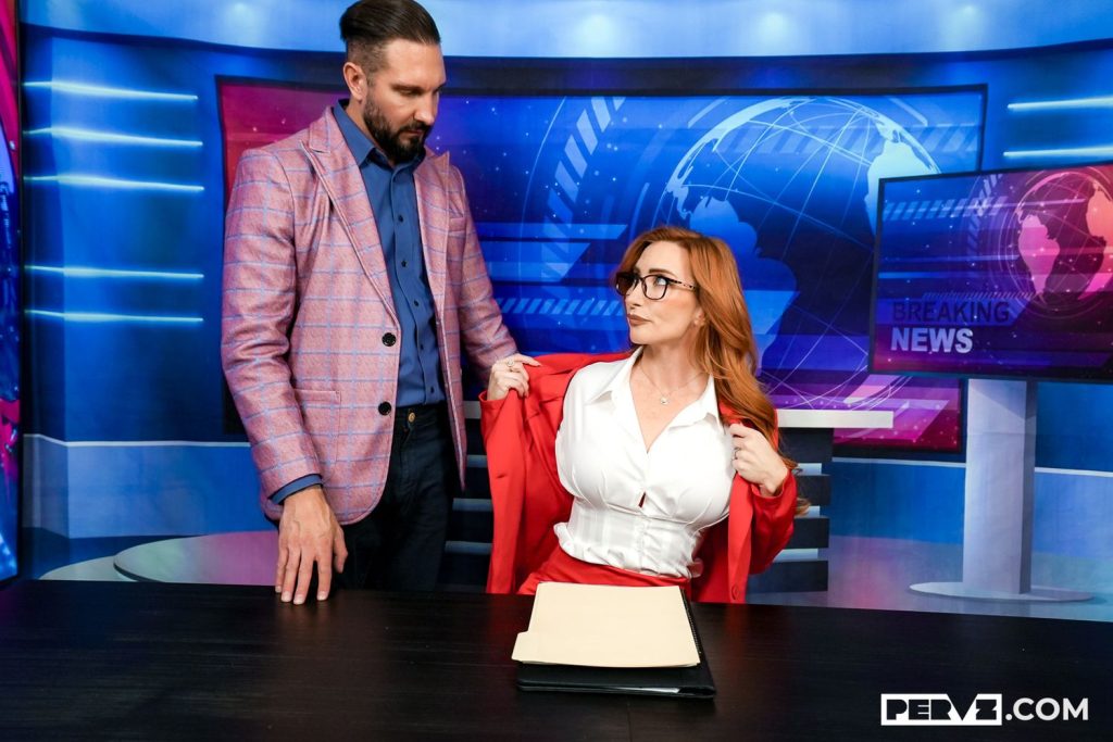 A man in a pink blazer stands next to a seated woman with red hair and glasses, who is adjusting her red jacket. They are in a news studio setting with Breaking News graphics on screens. A folder lies on the desk.