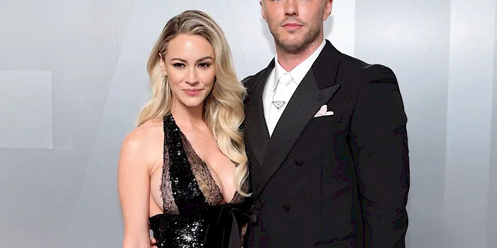Cropped photo of a man and a woman stand close together at a red carpet event. The woman wears a sleek black dress and the man wears a suit.