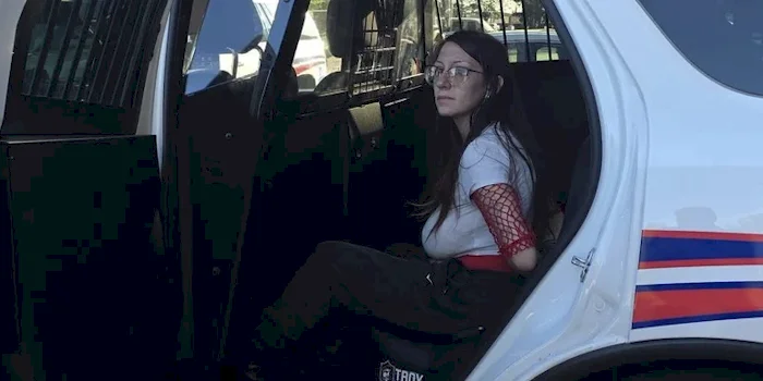 A woman sitting in the back of a police car. She is wearing glasses and her breasts look very large in a t-shirt